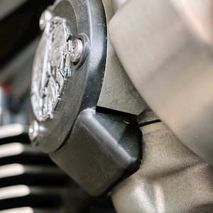 May include: A close-up of a motorcycle engine component featuring a circular black cover with a silver metallic emblem in the center. The assembly includes a smooth black housing, exposed silver bolts, and a textured metallic engine block surface.