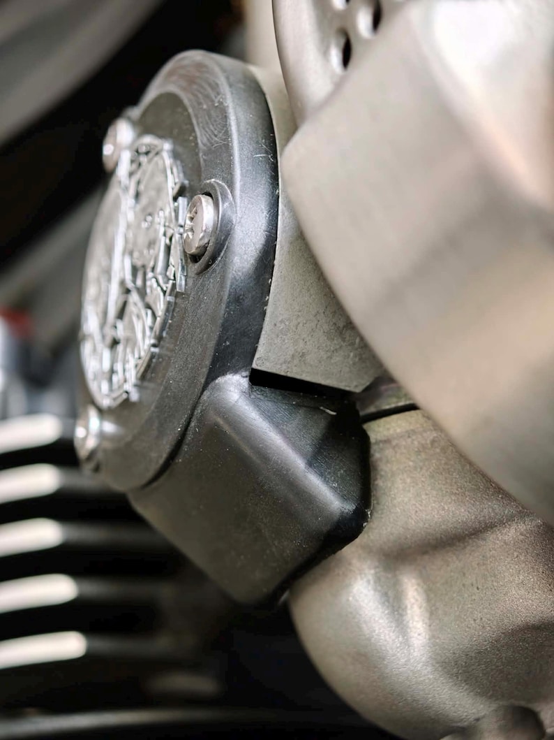 May include: A close-up of a motorcycle engine component featuring a circular black cover with a silver metallic emblem in the center. The assembly includes a smooth black housing, exposed silver bolts, and a textured metallic engine block surface.