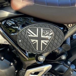 May include: Black motorcycle side panel with a Union Jack design and the number 77. The panel has a textured, diamond-patterned surface and is mounted on a black motorcycle frame. A brown leather seat is visible above the panel.