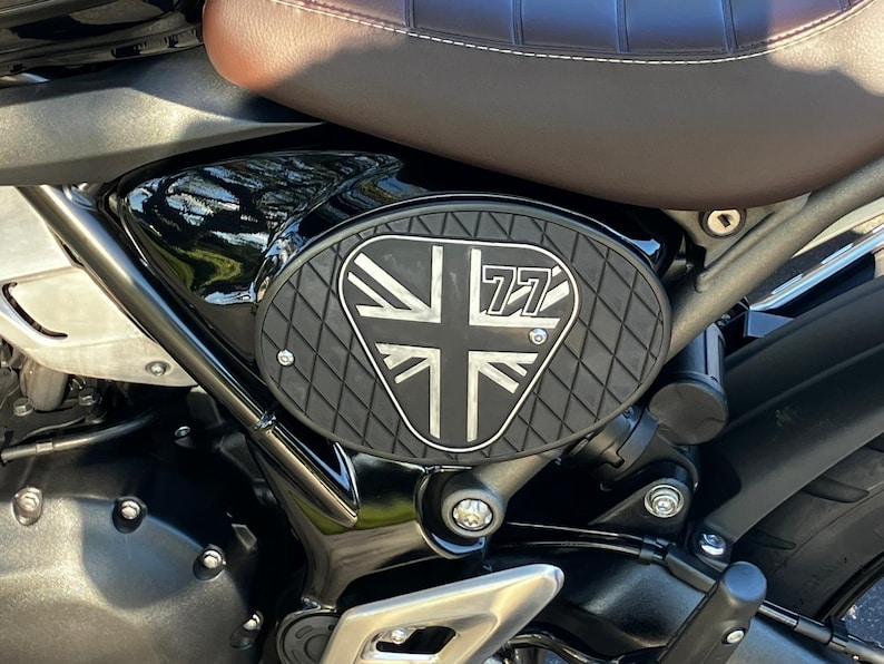 May include: Black motorcycle side panel with a Union Jack design and the number 77. The panel has a textured, diamond-patterned surface and is mounted on a black motorcycle frame. A brown leather seat is visible above the panel.