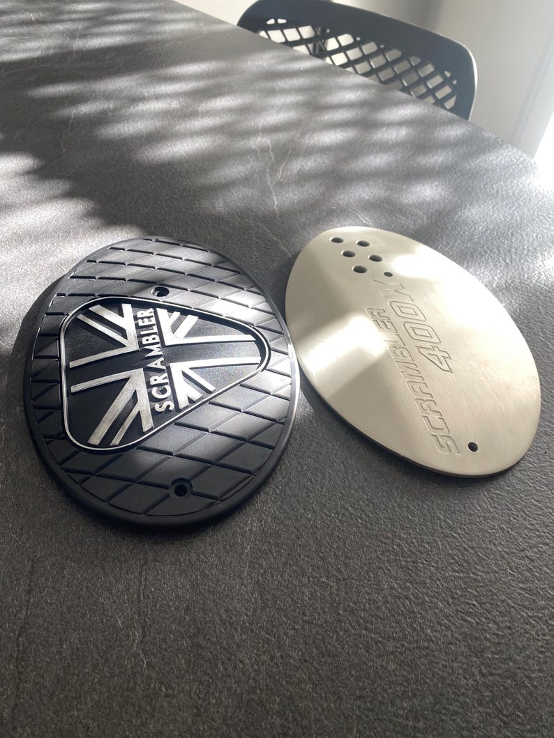May include: Two motorcycle parts. One is black with a diamond pattern and the Union Jack flag and the word "SCRAMBLER". The other is silver with the words "SCRAMBLER 400X".