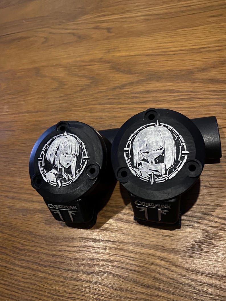 May include: Two black circular motorcycle brake reservoir covers featuring silver-toned engraved anime-style character portraits within a circular frame. Each cover displays the text CYBERPUNK and a logo at the bottom, with three mounting holes.