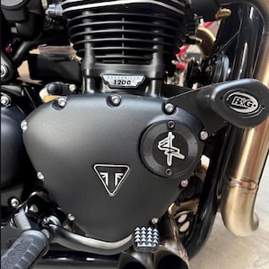 May include: A black motorcycle engine block featuring a Bonneville 1200 HT badge, a Triumph logo on the side cover, and an R&G crash protector. The engine includes metallic cooling fins, a circular textured cover with a white graphic, and dual exhaust pipes.