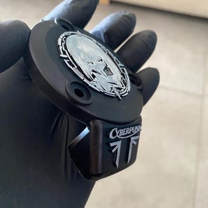 May include: A matte black circular emblem featuring a silver metallic inlay of a stylized character face within a geometric border. A rectangular extension below displays the silver text CYBERPUNK above two vertical silver geometric symbols.