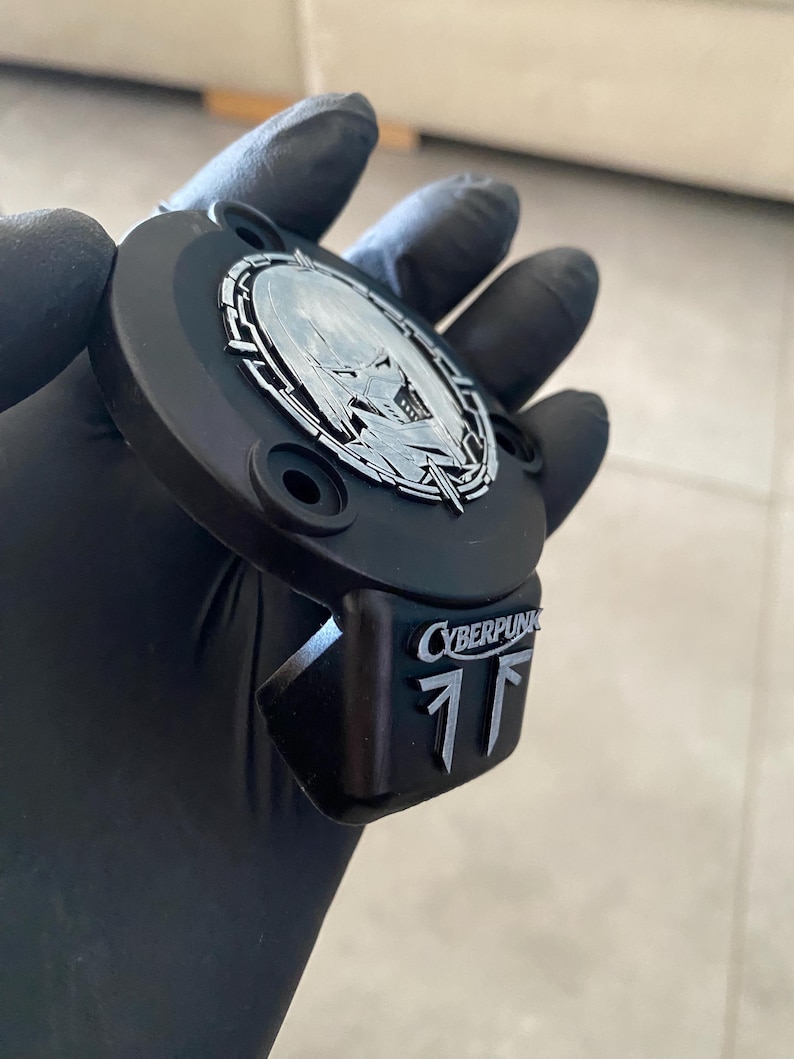 May include: A matte black circular emblem featuring a silver metallic inlay of a stylized character face within a geometric border. A rectangular extension below displays the silver text CYBERPUNK above two vertical silver geometric symbols.