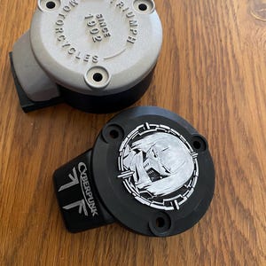 May include: Two circular motorcycle reservoir covers. One is silver with the embossed text TRIUMPH MOTORCYCLES SINCE 1902. The other is black with a silver circular graphic of a stylized character face and the text CYBERPUNK printed on the side.