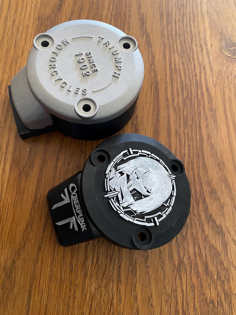 May include: Two circular motorcycle reservoir covers. One is silver with the embossed text TRIUMPH MOTORCYCLES SINCE 1902. The other is black with a silver circular graphic of a stylized character face and the text CYBERPUNK printed on the side.