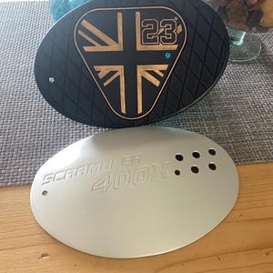 May include: Black and gold oval-shaped motorcycle part with a Union Jack design and the number 23. Below it, a silver oval part with the text "SCRAMBLER 400X" and several holes. The parts are on a wooden surface.