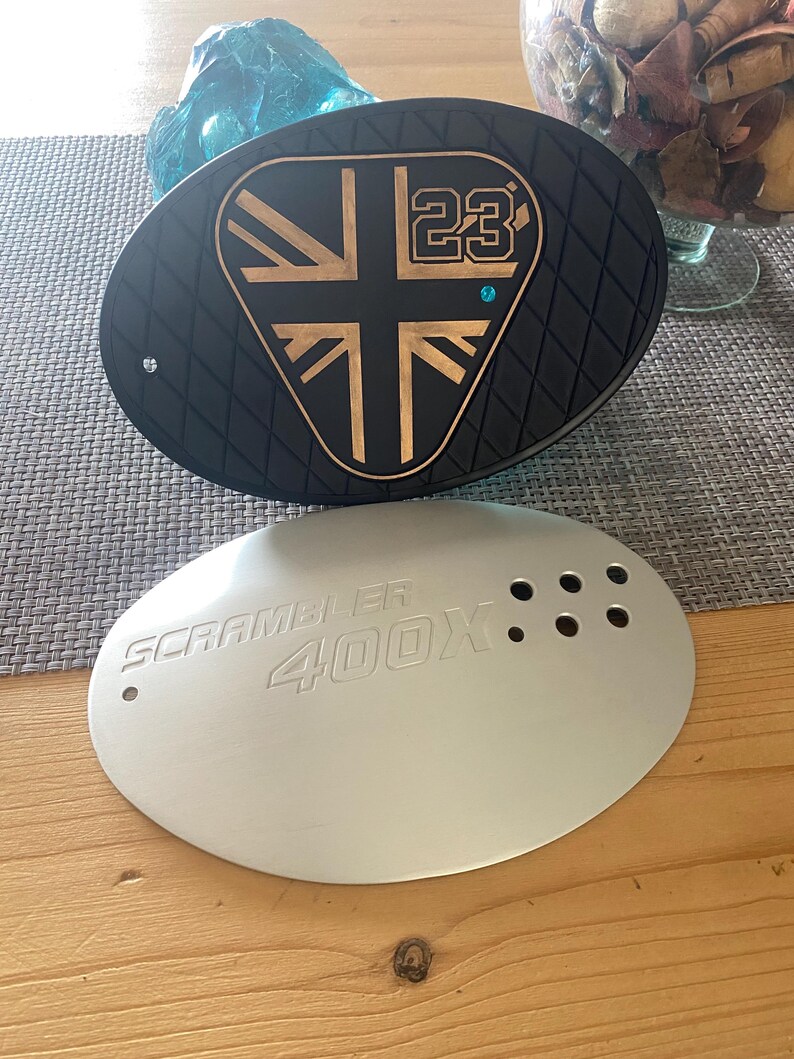 May include: Black and gold oval-shaped motorcycle part with a Union Jack design and the number 23. Below it, a silver oval part with the text "SCRAMBLER 400X" and several holes. The parts are on a wooden surface.