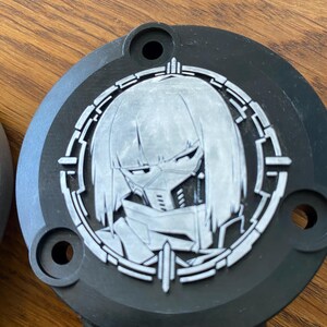 May include: A circular black object featuring a silver-toned relief of a stylized character with short hair and a face mask, enclosed within a geometric circular frame. The word CYBERPUNK is embossed in silver lettering at the bottom of the piece.