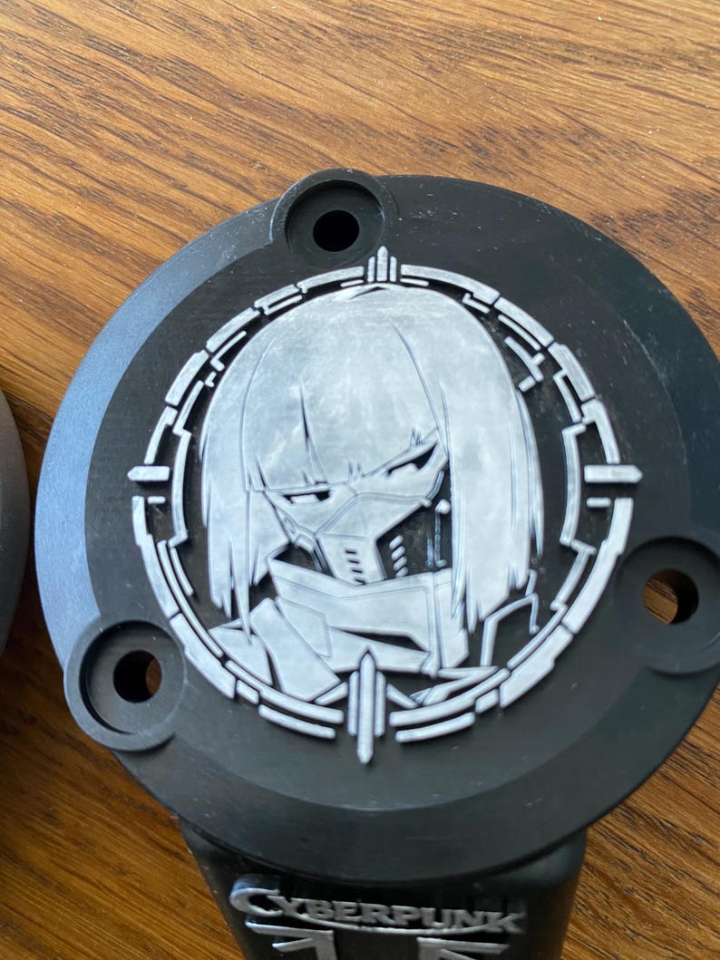 May include: A circular black object featuring a silver-toned relief of a stylized character with short hair and a face mask, enclosed within a geometric circular frame. The word CYBERPUNK is embossed in silver lettering at the bottom of the piece.