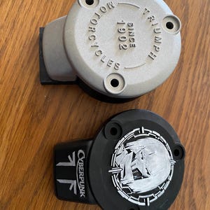 May include: Two circular motorcycle reservoir covers. One is silver with the embossed text "TRIUMPH MOTORCYCLES SINCE 1902" and three mounting holes. The other is black with a white graphic of a stylized character and the text "CYBERPUNK".