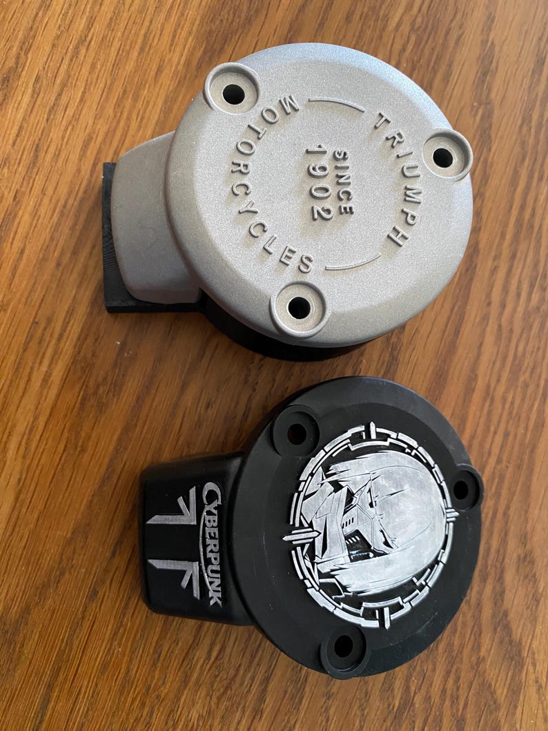 May include: Two circular motorcycle reservoir covers. One is silver with the embossed text "TRIUMPH MOTORCYCLES SINCE 1902" and three mounting holes. The other is black with a white graphic of a stylized character and the text "CYBERPUNK".