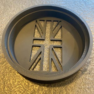 K&ouml;nnte beinhalten: Ein schwarzer, kreisf&ouml;rmiger Untersetzer mit einem ausgeschnittenen Union Jack-Design. Der Untersetzer hat einen erh&ouml;hten Rand und ein detailliertes Flaggenmuster in der Mitte. Der Durchmesser betr&auml;gt ca. 10 cm.