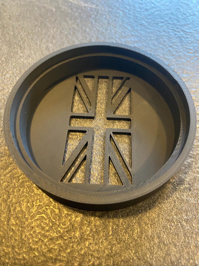 K&ouml;nnte beinhalten: Ein schwarzer, kreisf&ouml;rmiger Untersetzer mit einem ausgeschnittenen Union Jack-Design. Der Untersetzer hat einen erh&ouml;hten Rand und ein detailliertes Flaggenmuster in der Mitte. Der Durchmesser betr&auml;gt ca. 10 cm.