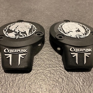 May include: Two matte black circular covers featuring a silver-toned anime-style character portrait within a circular frame. Below the portrait, each piece displays the word CYBERPUNK in silver lettering above a stylized silver geometric symbol.