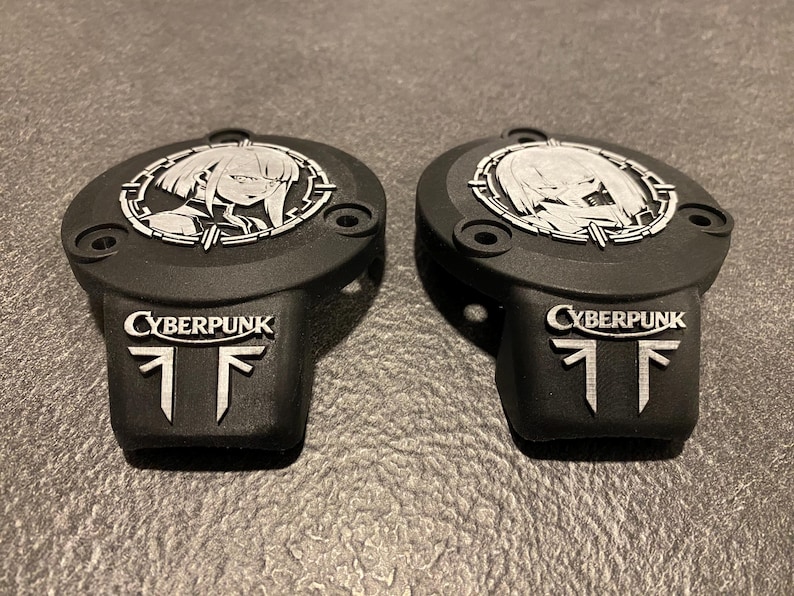 May include: Two matte black circular covers featuring a silver-toned anime-style character portrait within a circular frame. Below the portrait, each piece displays the word CYBERPUNK in silver lettering above a stylized silver geometric symbol.