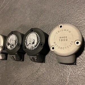 May include: Four circular motorcycle brake reservoir covers arranged in a row. Two are silver with the text TRIUMPH SINCE 1902 MOTORCYCLES, and two are black with a white graphic of a stylized character and the text CYBERPUNK on the lower tab.