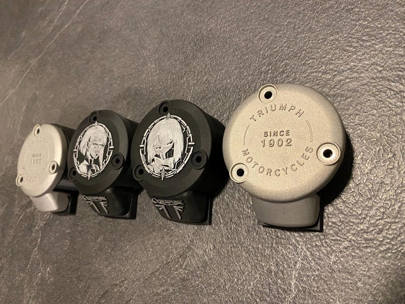 May include: Four circular motorcycle brake reservoir covers arranged in a row. Two are silver with the text TRIUMPH SINCE 1902 MOTORCYCLES, and two are black with a white graphic of a stylized character and the text CYBERPUNK on the lower tab.