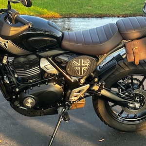 May include: Black motorcycle with a brown seat and a leather side bag. The motorcycle has a Union Jack emblem and gold-colored front suspension. The tires are black with a knobby tread.