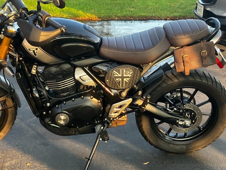 May include: Black motorcycle with a brown seat and a leather side bag. The motorcycle has a Union Jack emblem and gold-colored front suspension. The tires are black with a knobby tread.