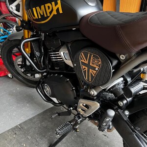 May include: Black motorcycle with gold accents, including the word "TRIUMPH" on the fuel tank. Features a brown seat and a side panel with a Union Jack design and the number 23. The motorcycle has a black engine and frame.