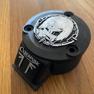 May include: A black circular mechanical component featuring a silver-colored emblem of a masked character within a circular frame on top. The side displays the text CYBERPUNK above a stylized logo. The object has three mounting holes on the top surface.