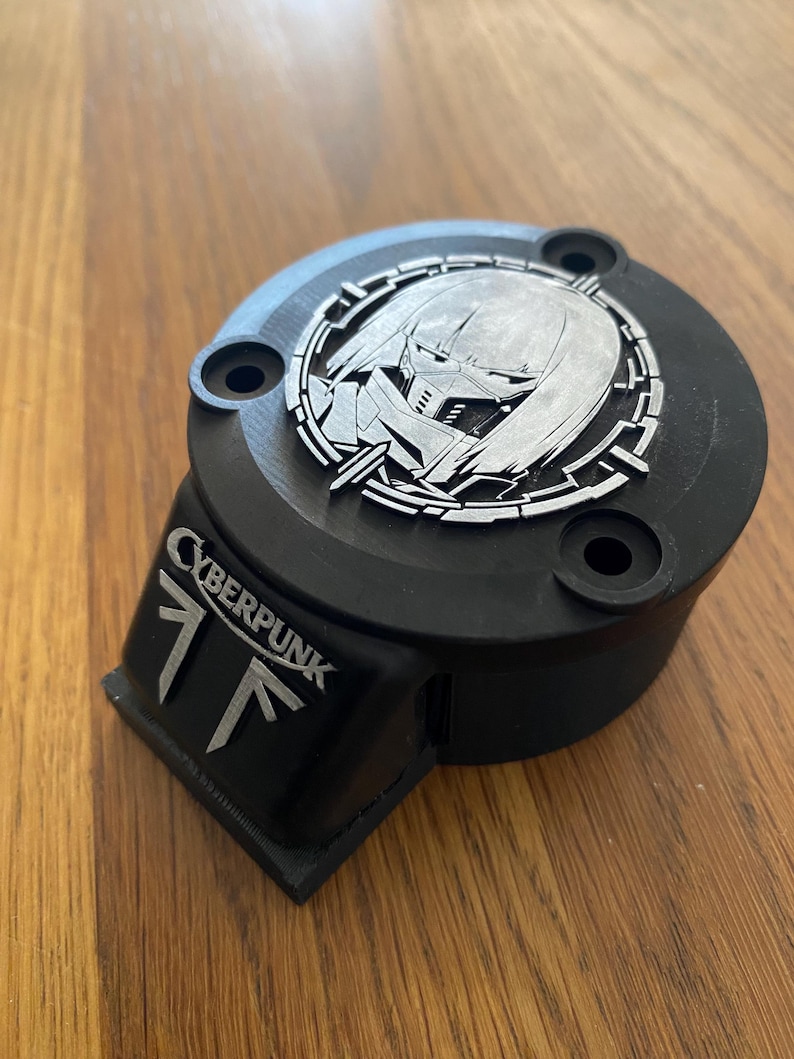 May include: A black circular mechanical component featuring a silver-colored emblem of a masked character within a circular frame on top. The side displays the text CYBERPUNK above a stylized logo. The object has three mounting holes on the top surface.