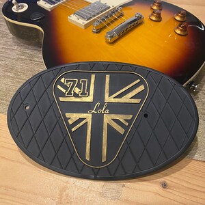 Może przedstawiać: Gitara elektryczna z wykończeniem sunburst spoczywa nad czarną, romboidalną plakietką. Plakietka zawiera złote litery i wz&oacute;r Union Jack z napisem "71 Lola".