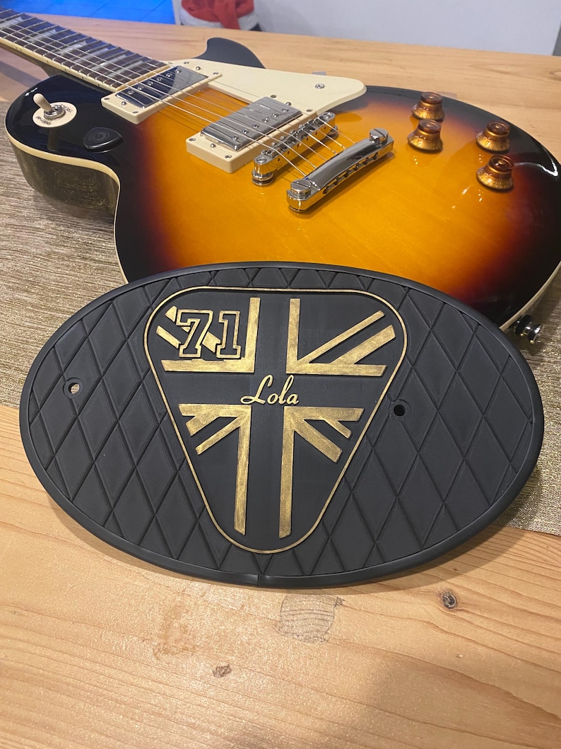 Może przedstawiać: Gitara elektryczna z wykończeniem sunburst spoczywa nad czarną, romboidalną plakietką. Plakietka zawiera złote litery i wz&oacute;r Union Jack z napisem "71 Lola".