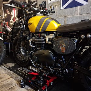 Może przedstawiać: Ż&oacute;łto-szary motocykl Triumph z czarnym siedzeniem i silnikiem. Zbiornik paliwa ma dwa szare paski. Motocykl ma czarną ramę i koła. Panel boczny przedstawia grafikę z kresk&oacute;wek. Motocykl jest zaparkowany w garażu.