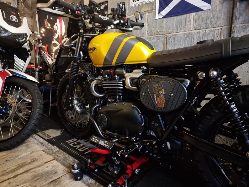 Może przedstawiać: Ż&oacute;łto-szary motocykl Triumph z czarnym siedzeniem i silnikiem. Zbiornik paliwa ma dwa szare paski. Motocykl ma czarną ramę i koła. Panel boczny przedstawia grafikę z kresk&oacute;wek. Motocykl jest zaparkowany w garażu.