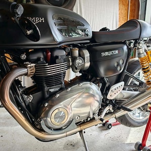 May include: A matte black Triumph Thruxton R motorcycle featuring a fuel tank with a Union Jack emblem, a black seat with red stitching, silver engine casing with a gold Triumph logo, and yellow rear suspension springs on a red paddock stand.