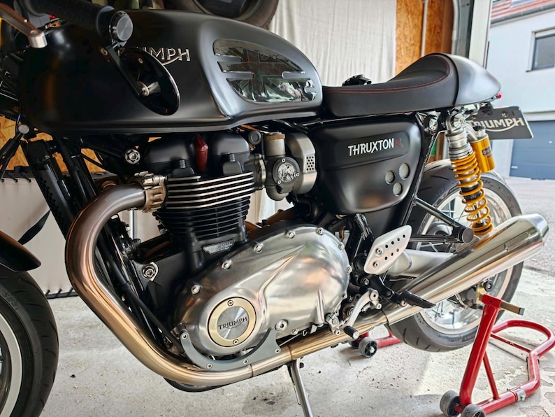 May include: A matte black Triumph Thruxton R motorcycle featuring a fuel tank with a Union Jack emblem, a black seat with red stitching, silver engine casing with a gold Triumph logo, and yellow rear suspension springs on a red paddock stand.