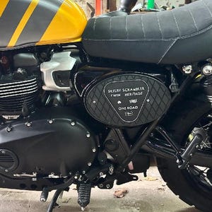 Może przedstawiać: Czarno-ż&oacute;łty motocykl z czarnym siedzeniem i oponami. Zbiornik paliwa jest ż&oacute;łty z szarymi paskami i napisem "Triumph". Na panelu bocznym widnieje napis "SKELBY SCRAMBLER TWIN HERITAGE ONE ROAD".
