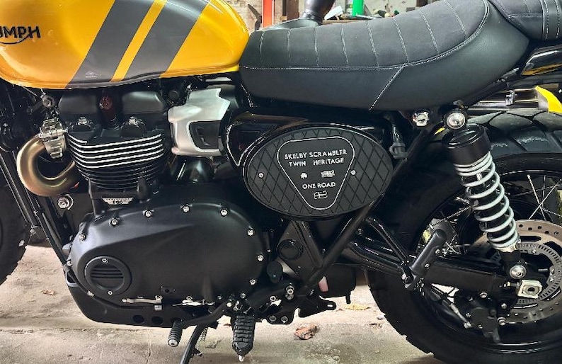 Może przedstawiać: Czarno-ż&oacute;łty motocykl z czarnym siedzeniem i oponami. Zbiornik paliwa jest ż&oacute;łty z szarymi paskami i napisem "Triumph". Na panelu bocznym widnieje napis "SKELBY SCRAMBLER TWIN HERITAGE ONE ROAD".