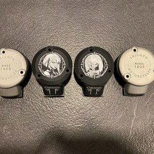 May include: Four circular motorcycle reservoir covers arranged in a row. Two silver covers feature the text Triumph Motorcycles Since 1902. Two black covers feature white anime-style character illustrations and the text Cyberpunk at the base.