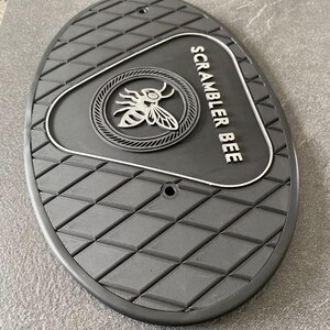 Może przedstawiać: Czarny, owalny podn&oacute;żek motocyklowy z rombowym wzorem. Posiada okrągły emblemat z motywem pszczoły i biały napis "SCRAMBLER BEE". Dwa małe otwory są widoczne w g&oacute;rnej części.