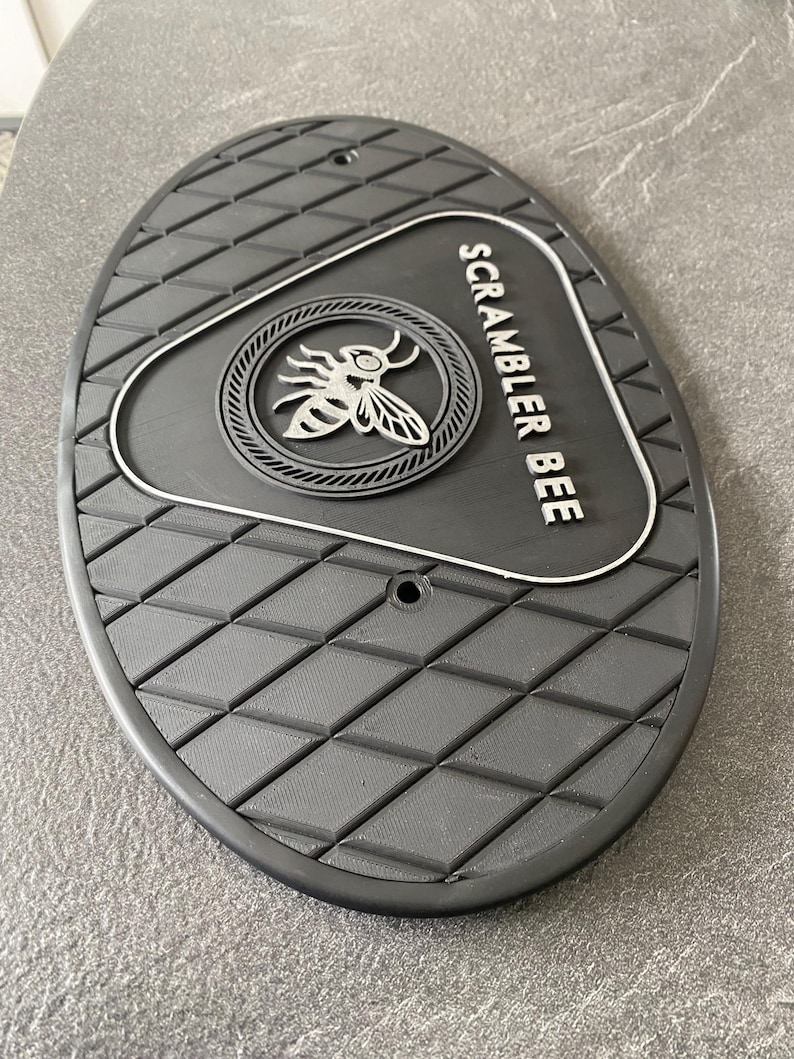 Może przedstawiać: Czarny, owalny podn&oacute;żek motocyklowy z rombowym wzorem. Posiada okrągły emblemat z motywem pszczoły i biały napis "SCRAMBLER BEE". Dwa małe otwory są widoczne w g&oacute;rnej części.