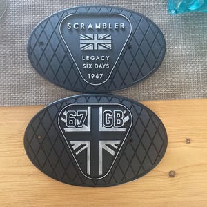 Może przedstawiać: Dwie czarne, owalne metalowe tabliczki z rombowym tłem. Na g&oacute;rnej tabliczce napis "SCRAMBLER" nad Union Jack i "LEGACY SIX DAYS 1967". Dolna tabliczka ma "67 GB" i wz&oacute;r Union Jack.