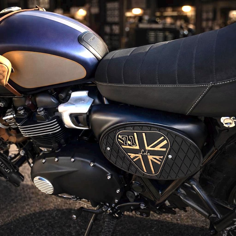 Może przedstawiać: Niestandardowy motocykl z niebieskim i beżowym zbiornikiem paliwa, czarnym siedzeniem i panelem bocznym. Panel boczny ma wz&oacute;r Union Jack z napisem "71 Lola". Motocykl ma klasyczną, zabytkową estetykę.