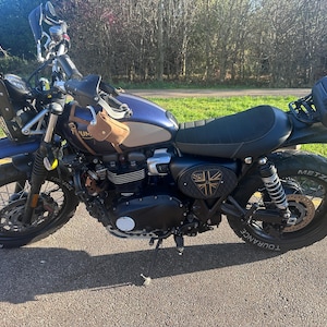 Może przedstawiać: Niebiesko-srebrny motocykl z czarnymi oponami i czarnym siedzeniem. Motocykl ma małą brązową torbę przymocowaną z przodu i czarną torbę do przechowywania z tyłu. Motocykl ma emblemat Union Jack z boku.