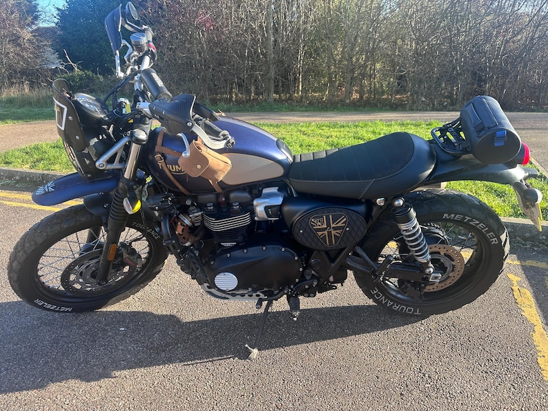 Może przedstawiać: Niebiesko-srebrny motocykl z czarnymi oponami i czarnym siedzeniem. Motocykl ma małą brązową torbę przymocowaną z przodu i czarną torbę do przechowywania z tyłu. Motocykl ma emblemat Union Jack z boku.