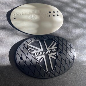 May include: Two oval-shaped motorcycle parts. One is light gray with the text "SCRAMBLER 400X". The other is black with a diamond pattern and a Union Jack design with the word "SCRAMBLER".