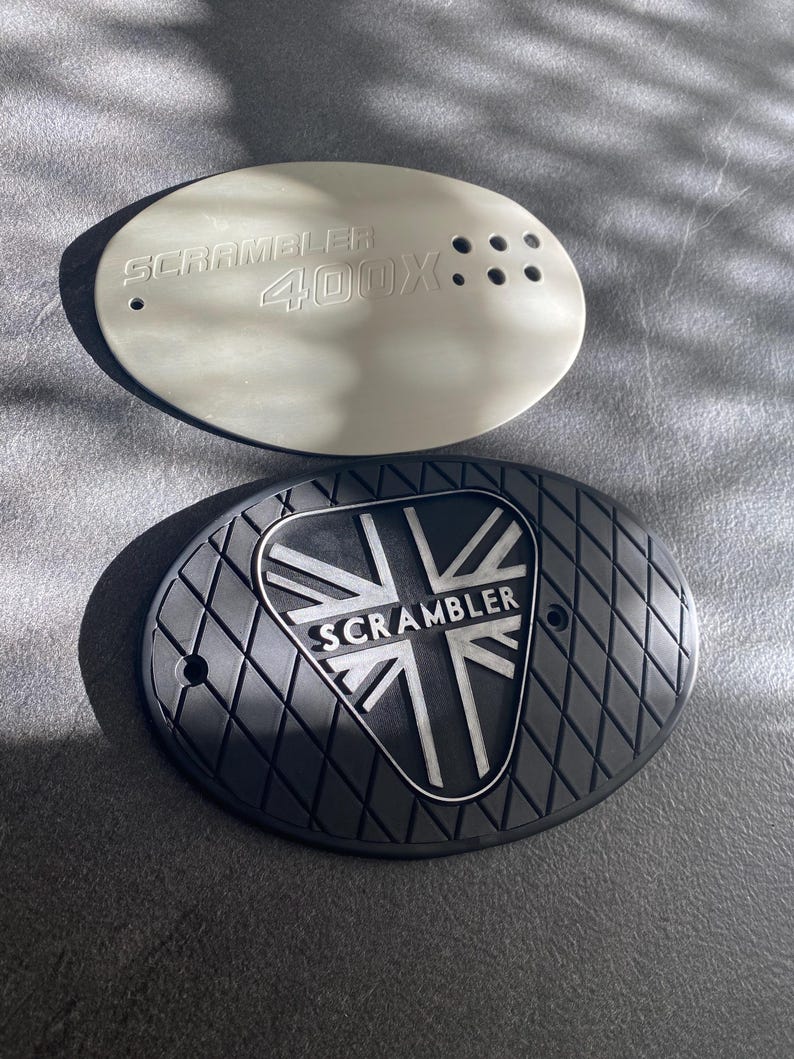 May include: Two oval-shaped motorcycle parts. One is light gray with the text "SCRAMBLER 400X". The other is black with a diamond pattern and a Union Jack design with the word "SCRAMBLER".