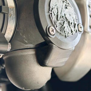 May include: A matte black automotive engine component featuring a circular emblem with an engraved anime-style character design. The part includes a metal clamp with the text MPC 1241705 visible on the side, secured by silver-toned screws.
