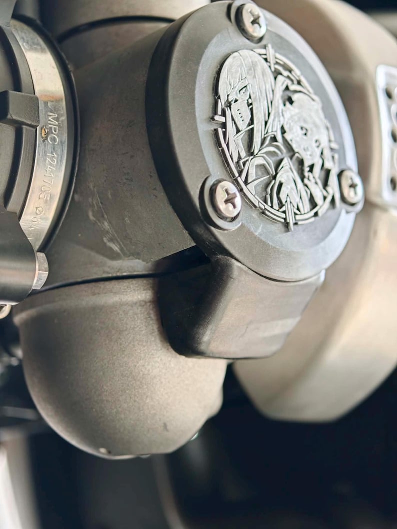 May include: A matte black automotive engine component featuring a circular emblem with an engraved anime-style character design. The part includes a metal clamp with the text MPC 1241705 visible on the side, secured by silver-toned screws.