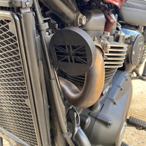 K&ouml;nnte beinhalten: Nahaufnahme eines Motorradmotors mit einer schwarzen Auspuffabdeckung mit Union Jack-Design. Die Motorbestandteile umfassen einen K&uuml;hler, ein Auspuffrohr und verschiedene mechanische Teile. Das Motorrad hat einen rot-wei&szlig;en Tank.
