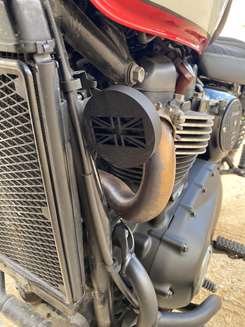 K&ouml;nnte beinhalten: Nahaufnahme eines Motorradmotors mit einer schwarzen Auspuffabdeckung mit Union Jack-Design. Die Motorbestandteile umfassen einen K&uuml;hler, ein Auspuffrohr und verschiedene mechanische Teile. Das Motorrad hat einen rot-wei&szlig;en Tank.