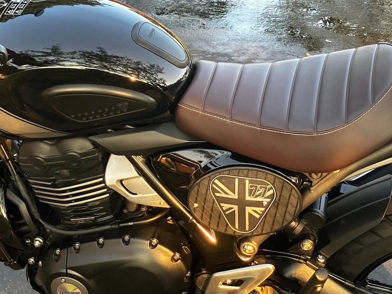 May include: Close-up of a black motorcycle with a brown leather seat. The motorcycle has a Union Jack emblem with the number 77 on the side. The motorcycle is parked outside.